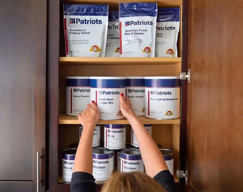 A person organizes a pantry shelf stocked with 4Patriots emergency food supplies, including cans of chicken, pork, beef, black bean burger mix, and freeze-dried blueberries, alongside pouches of mac and cheese, rice, and creamy wheat.