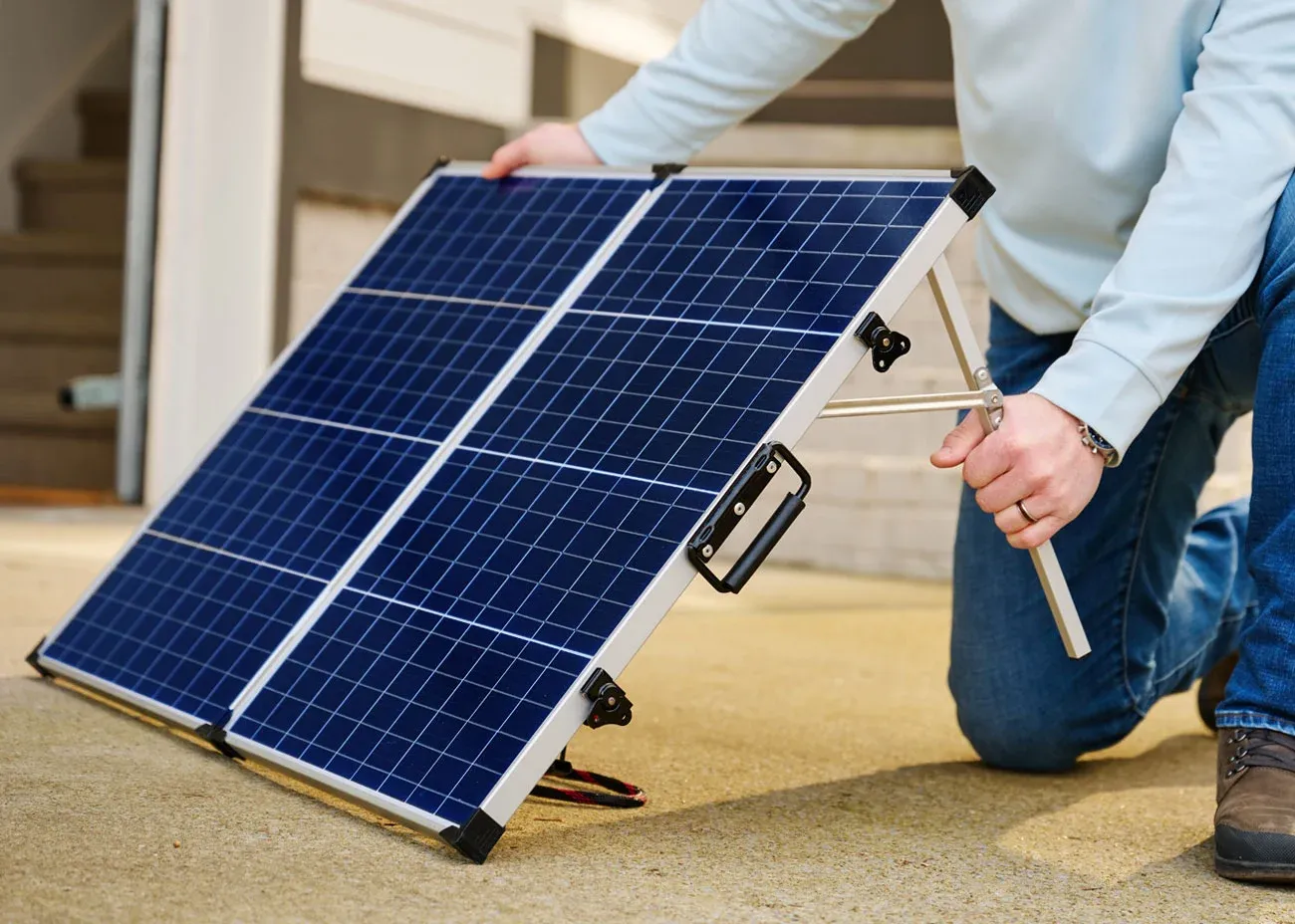 A person in jeans and a light blue sweatshirt kneels on the ground while setting up a portable 100-watt solar panel for a solar generator with a built-in stand. The panel is positioned outdoors on a cement surface, angled to capture sunlight for off-grid power generation.