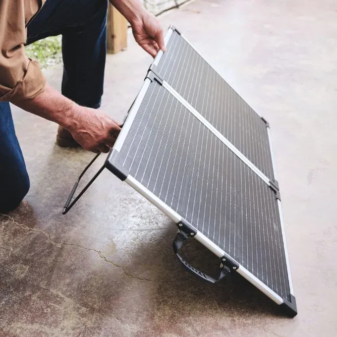 A person sets up a foldable portable solar panel on the ground, adjusting its angle for sunlight capture.