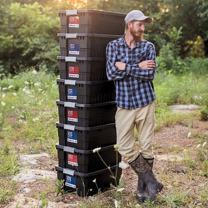 A man in a cap and boots stands beside a tall stack of 4Patriots 1-Year Survival Food Kit containers in a grassy field at sunset.