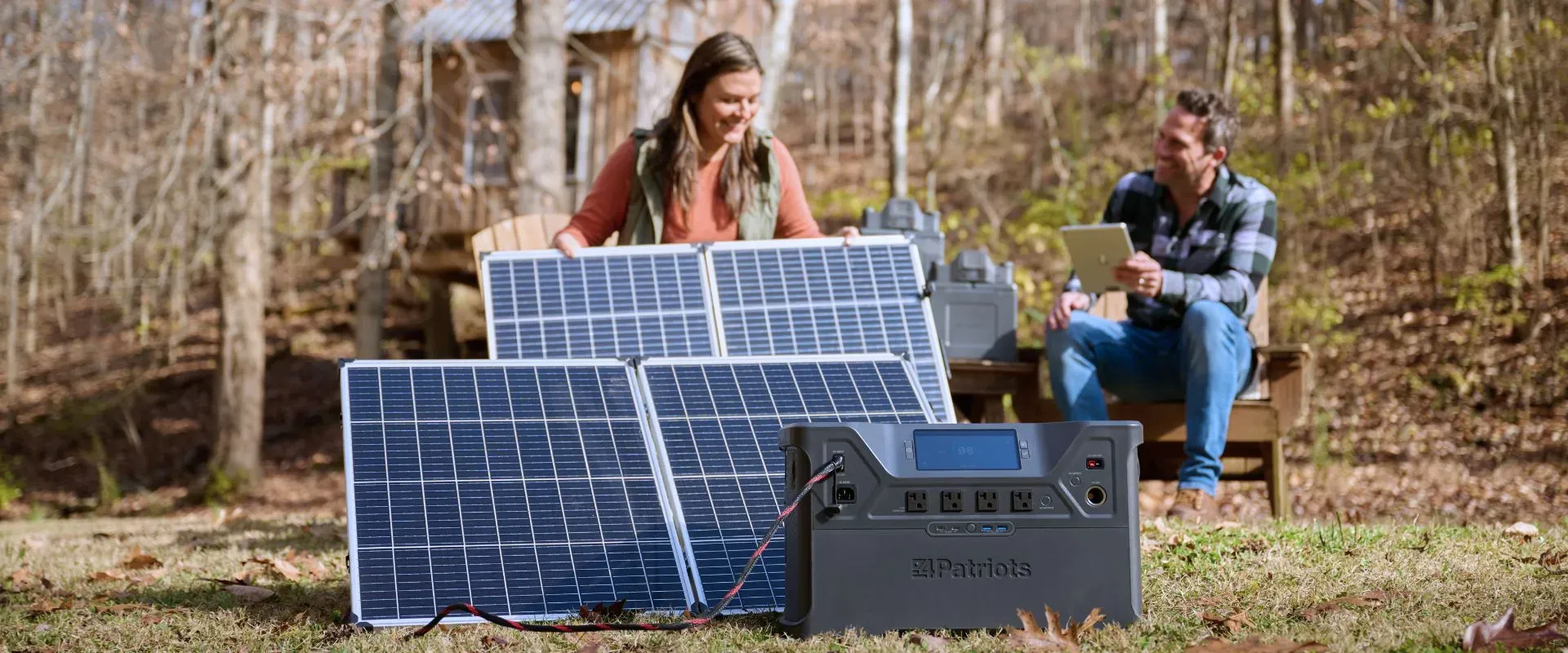A woman sets up portable solar panels on a grassy clearing while a man sits nearby on a wooden bench holding a tablet. In the foreground, a 4Patriots solar generator is connected to the panels, with autumn trees in the background.