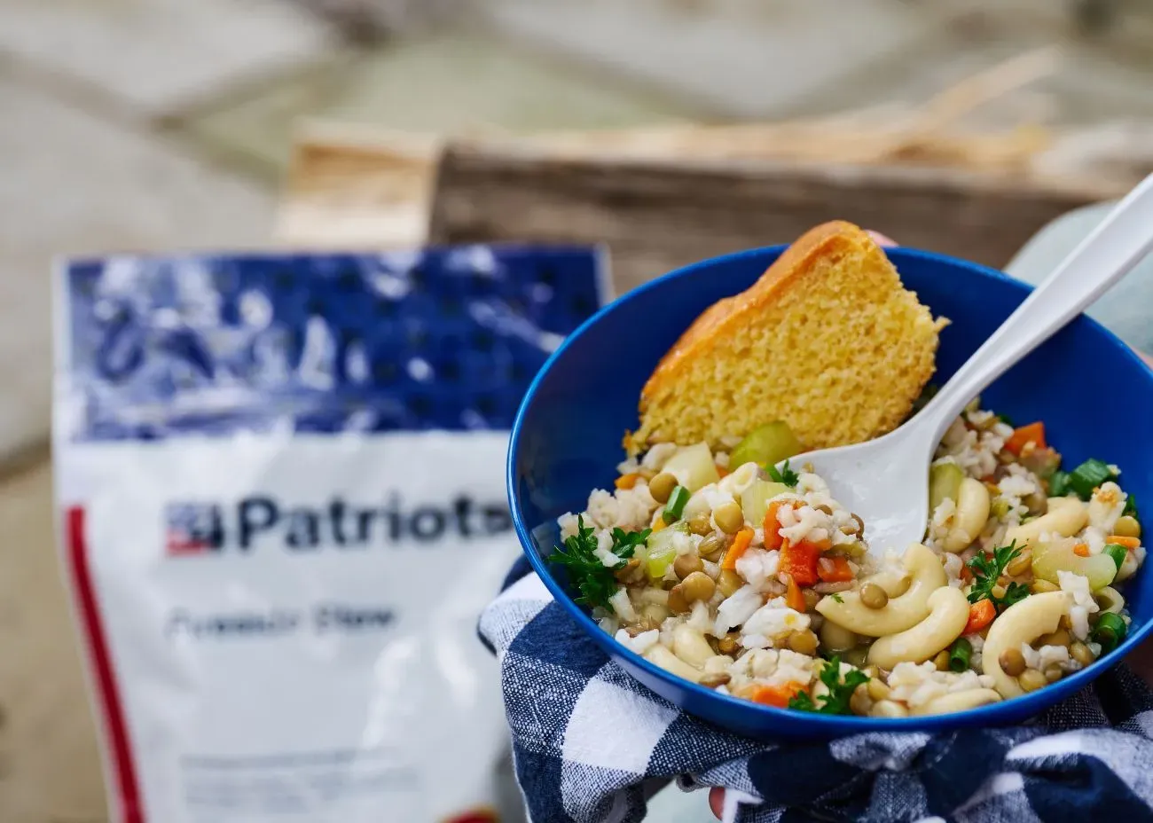 Close-up of a blue bowl filled with a hearty vegetable and pasta stew 4Patriots 4-Week Emergency Food Supply Kit featuring lentils, rice, macaroni, carrots, celery, and parsley, topped with a slice of cornbread. A white spoon rests in the bowl, and the background shows a blurred emergency food supply package labeled “Patriots” and “Freedom Stew.”