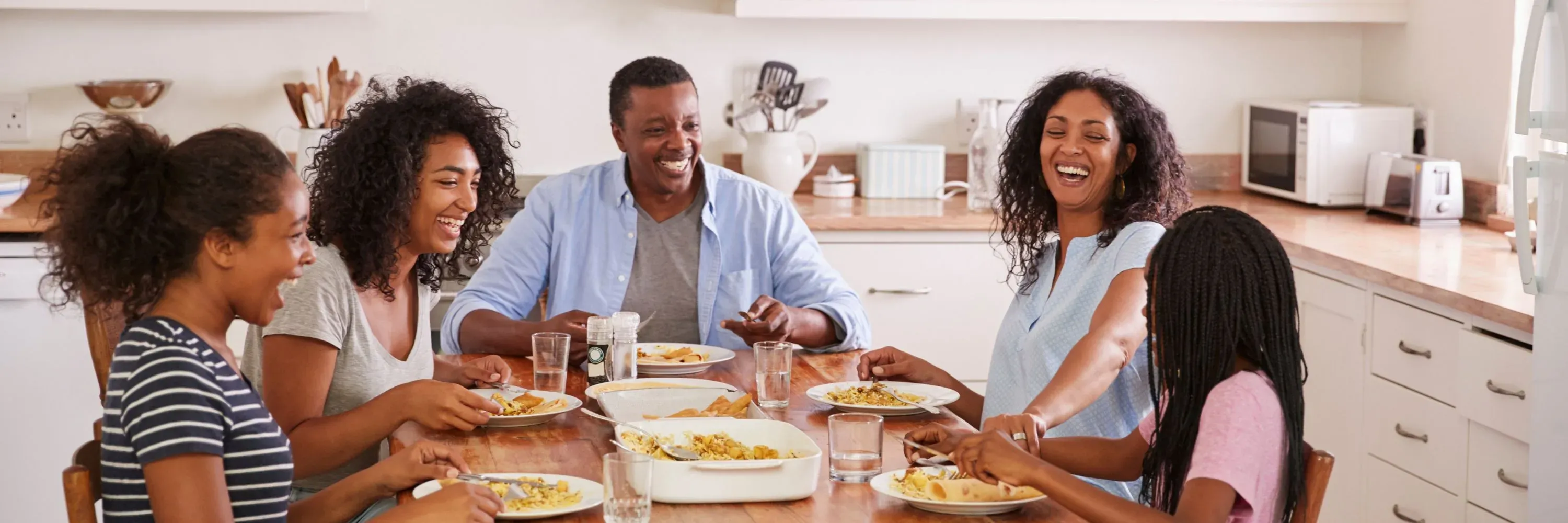 A smiling family gathered around a kitchen table enjoying a hot meal together, served from a casserole dish. The image emphasizes how an emergency food supply can support comforting, shared meals during uncertain times.