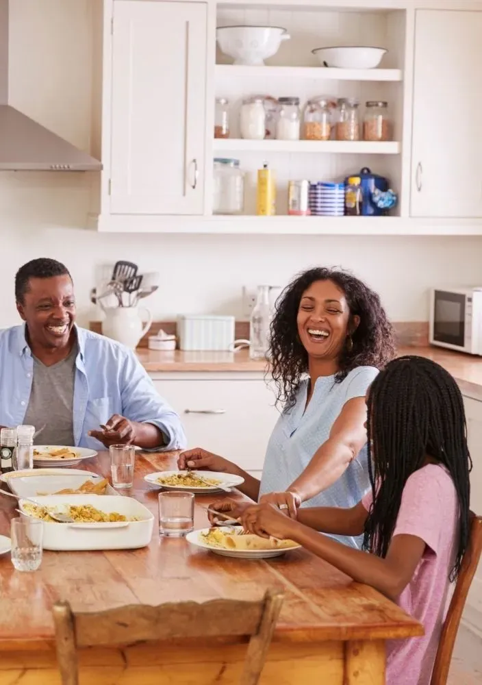 A smiling family gathered around a kitchen table enjoying a hot meal together, served from a casserole dish. The image emphasizes how an emergency food supply can support comforting, shared meals during uncertain times.