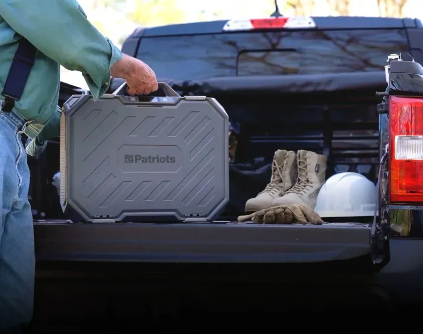 A person’s hand lifting a large, rugged grey 4Patriots AlphaCase portable power station from the bed of a black pickup truck.