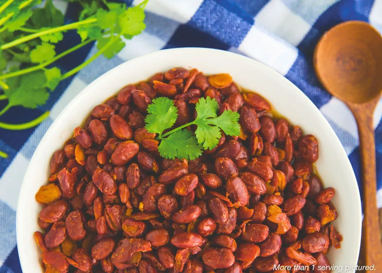 Authentic Red Beans Emergency Food Kit A white bowl of cooked authentic red beans garnished with a sprig of fresh cilantro, served on a blue and white checkered cloth next to a wooden spoon.