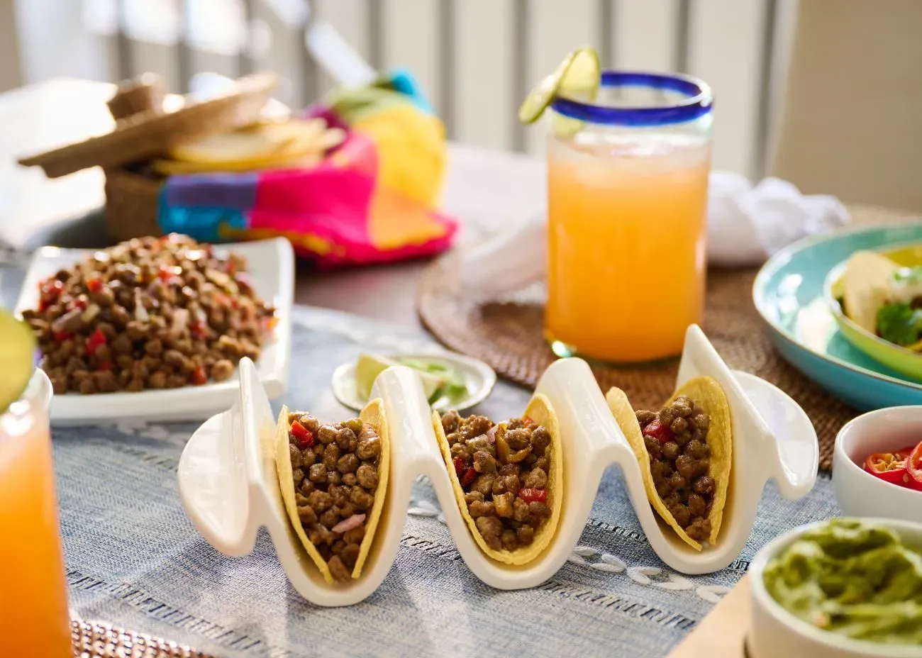 Three hard-shell tacos filled with beef-flavored plant-based protein bits and peppers, displayed in a white taco holder as part of a vibrant meal spread with guacamole and fruit drinks.