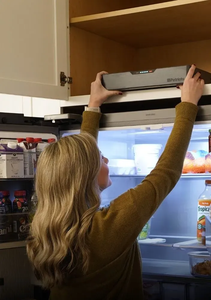 A woman reaching up to place a slim, dark grey 4Patriots Blackout Buddy Generator from a wooden cabinet shelf located directly above a stainless steel refrigerator.
