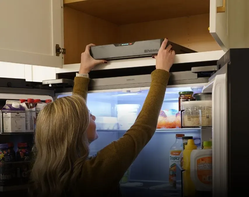 A woman reaching up to place a slim, dark grey 4Patriots Blackout Buddy Generator from a wooden cabinet shelf located directly above a stainless steel refrigerator.