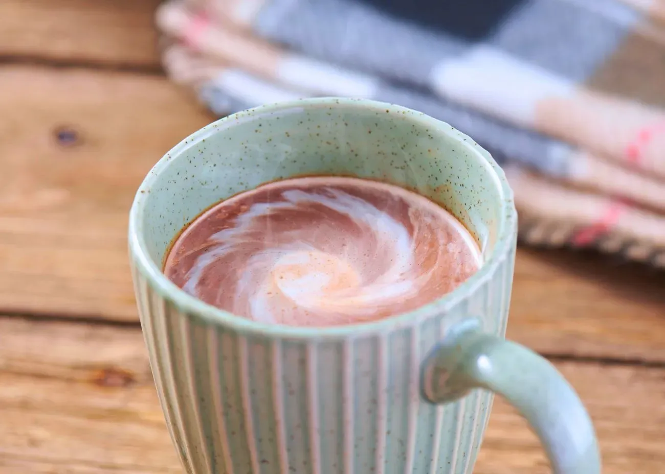 A steaming light-green ceramic mug of hot cocoa with a creamy white swirl on top, set against a rustic wooden background with a folded plaid blanket.