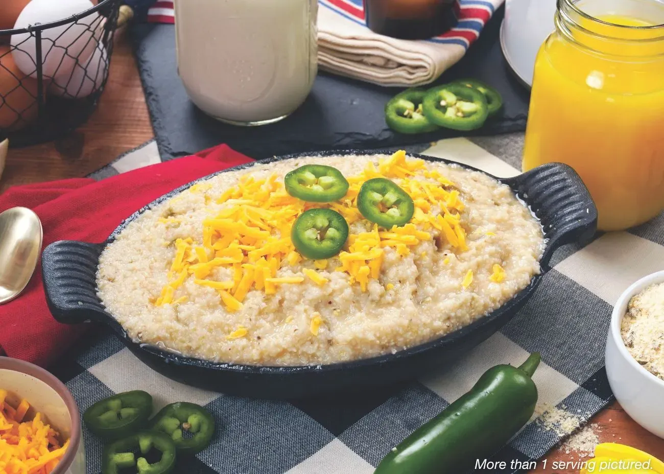 A black cast-iron serving dish filled with creamy green chili grits, topped with melted shredded cheddar cheese and fresh jalapeño slices, served on a black and white checkered cloth.