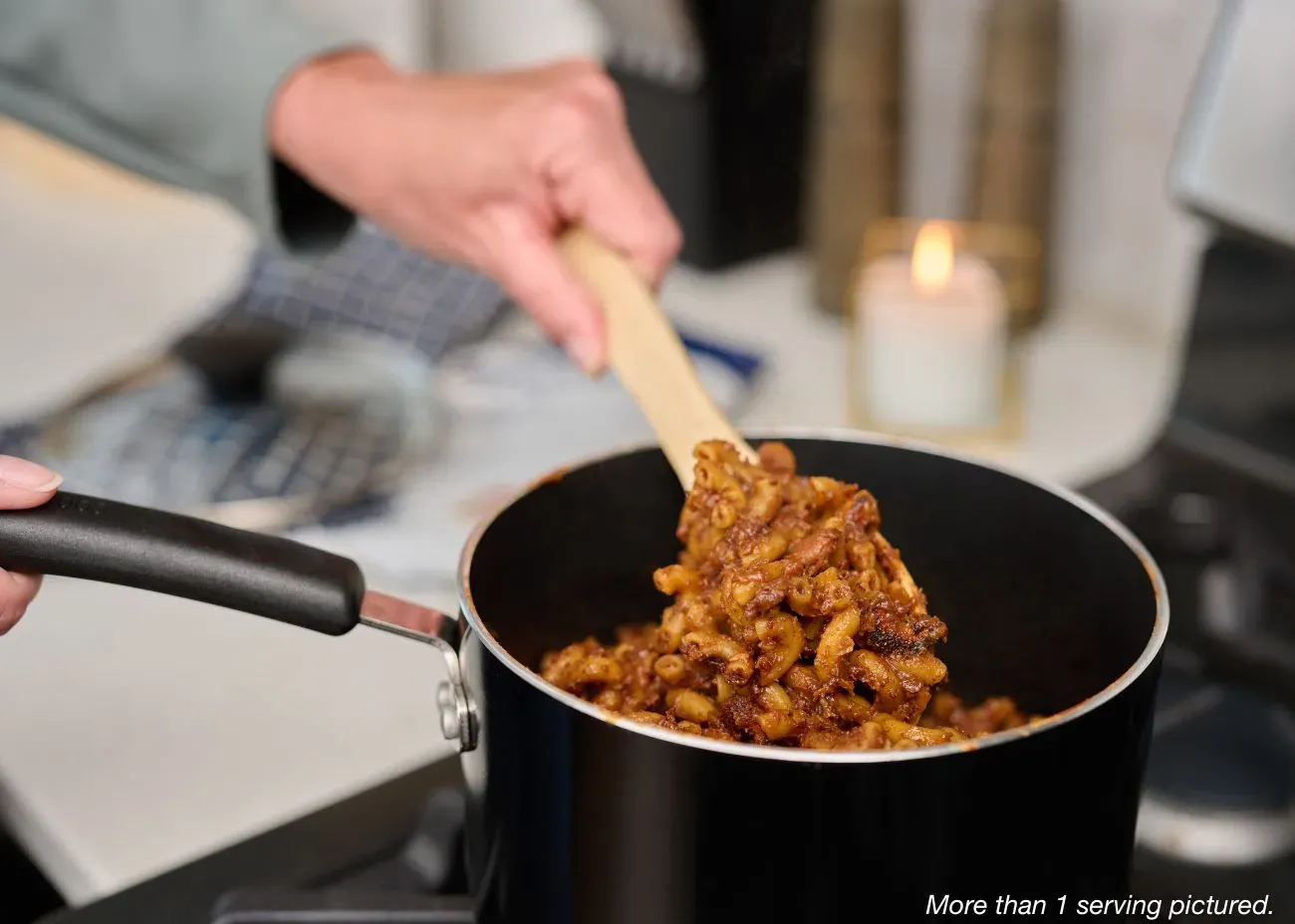 A close-up of a person using a wooden spoon to stir a steaming pot of hearty chili mac with macaroni noodles and meat sauce on a stovetop.