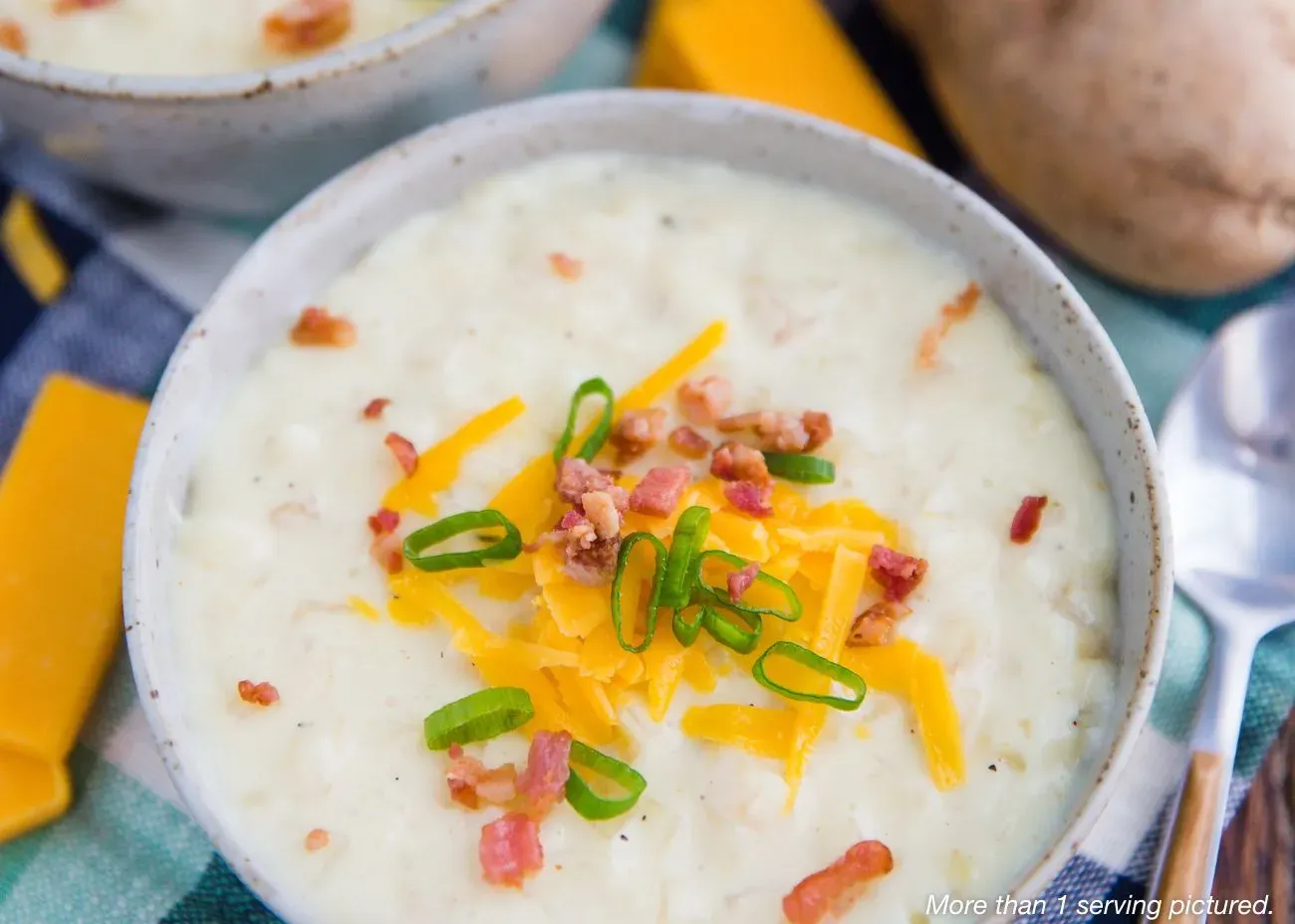 Cozy Potato Soup Emergency Food A close-up of a white bowl filled with thick, creamy potato soup topped with shredded cheddar cheese, crispy bacon bits, and sliced green onions, served on a green and white checkered cloth.