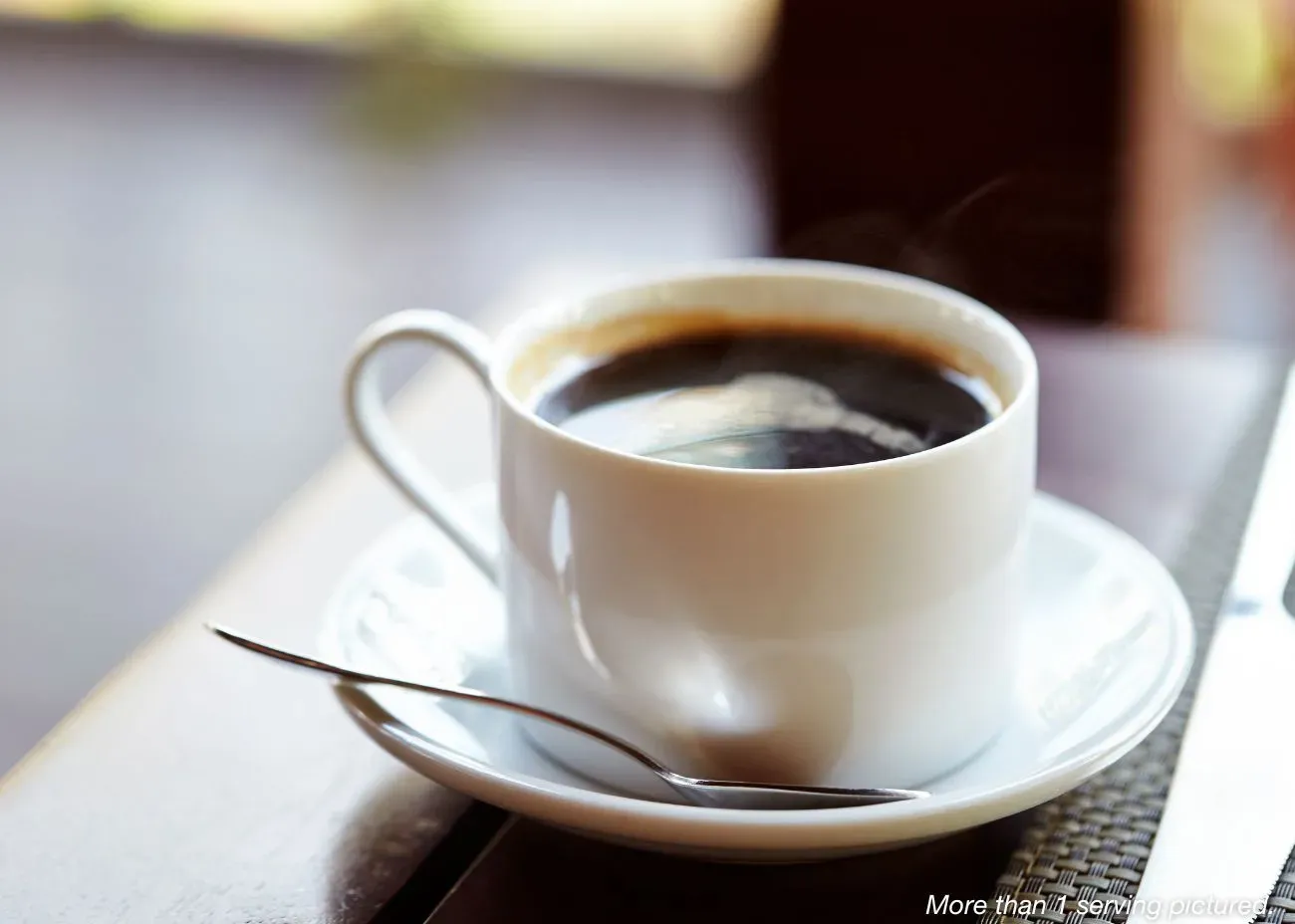 A steaming white ceramic mug of black coffee sitting on a matching saucer with a silver spoon, set on a dark wooden table in soft, natural light.