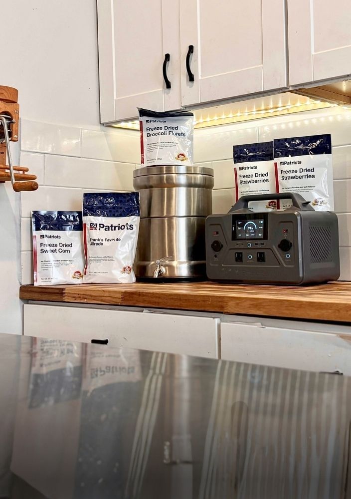A kitchen counter display featuring several bags of 4Patriots freeze-dried food (broccoli, strawberries, sweet corn, and alfredo), a large stainless steel water filtration system, and a portable solar power generator 1200.