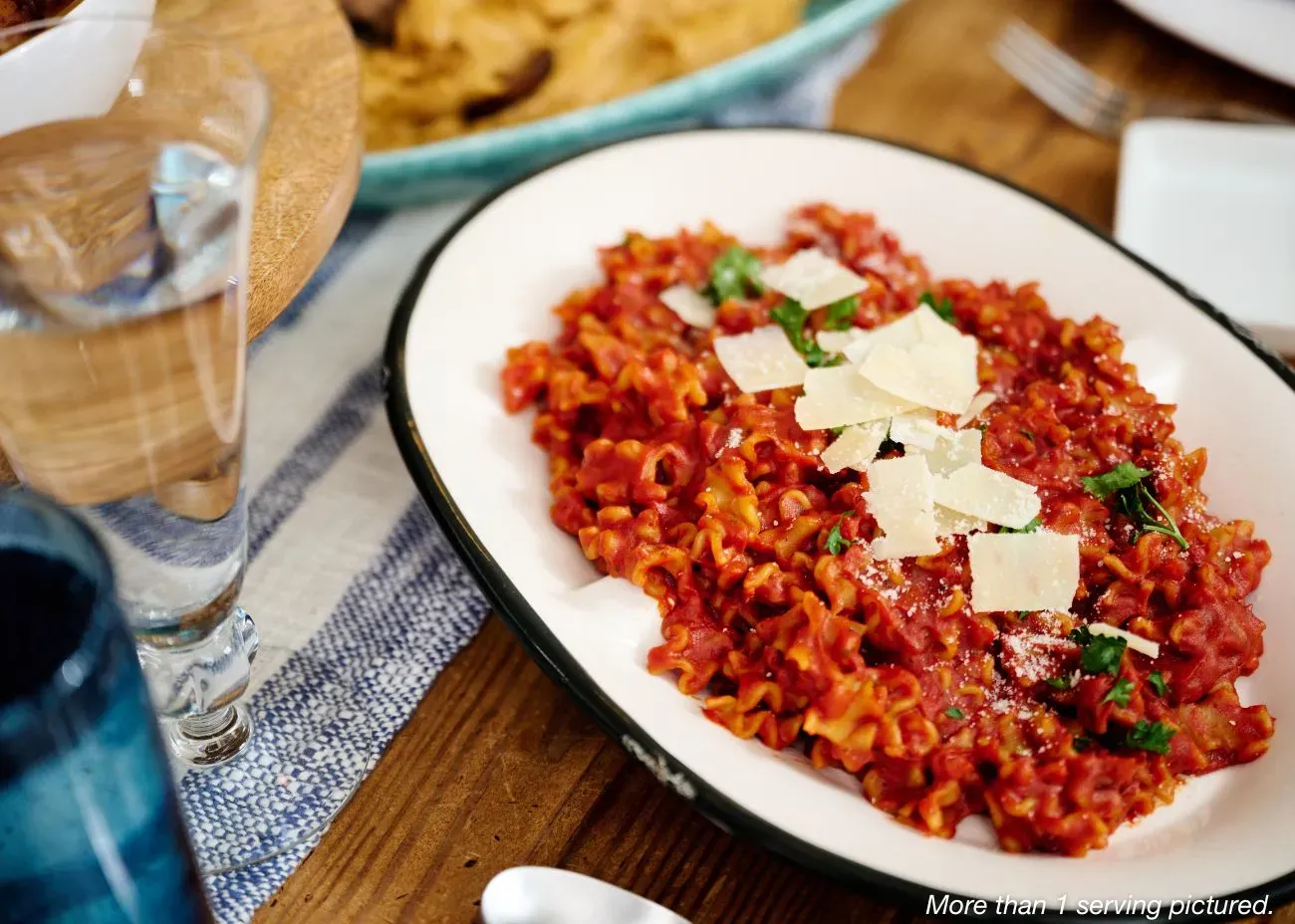 A white oval plate featuring pasta tossed in a vibrant red marinara sauce, topped with shaved parmesan cheese and fresh parsley.