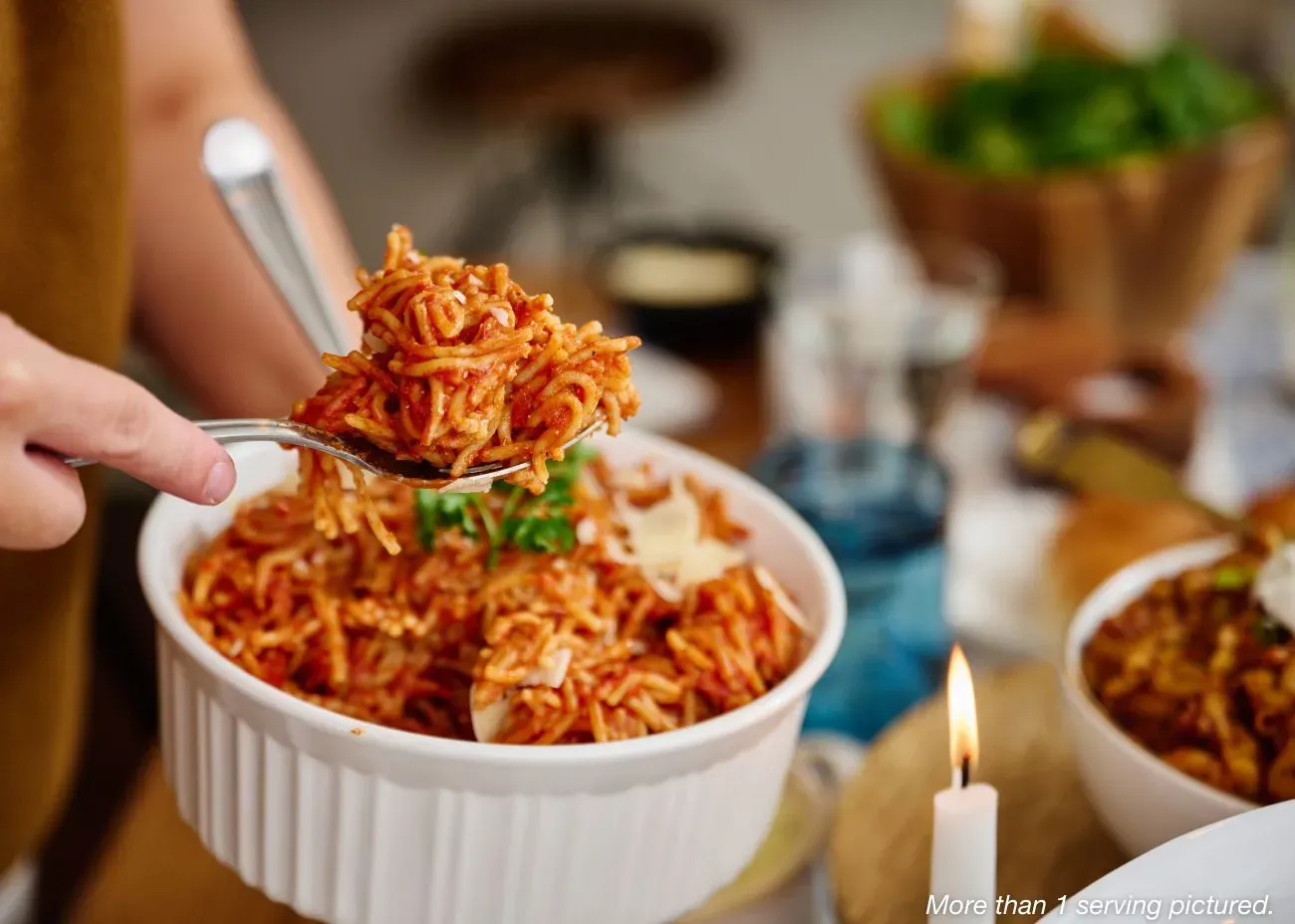 A large white serving bowl of spaghetti in red sauce; a person is shown lifting a large portion of the pasta with a fork and spoon.