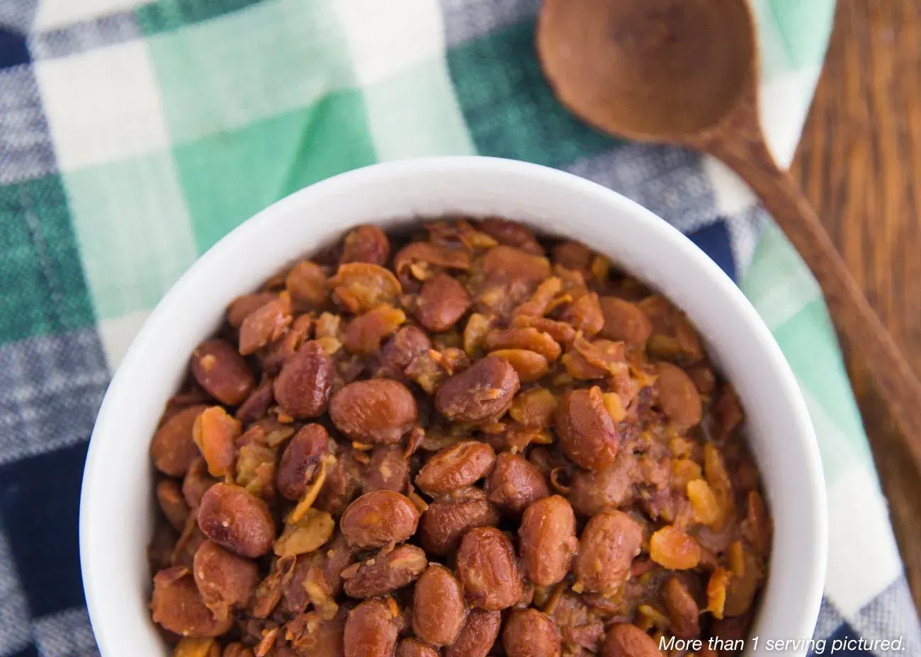 Southern Pinto Beans Emergency Food Kit A top-down view of a white bowl filled with cooked Southern pinto beans, served on a green and white checkered cloth next to a wooden spoon.