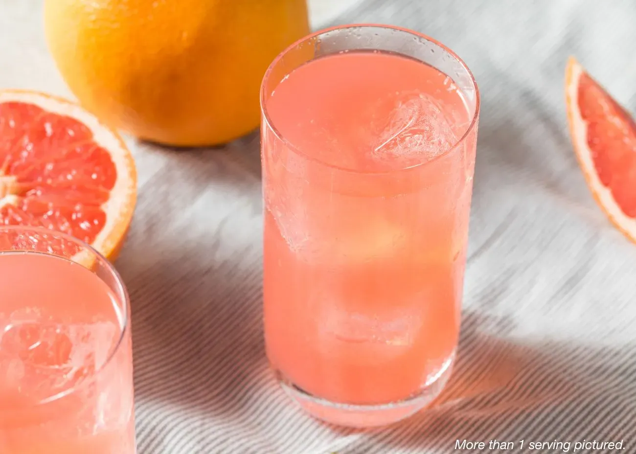 A close-up of a tall glass of pink-orange Star Spangled Tropical Energy Drink with ice, served on a striped cloth next to fresh grapefruit slices.