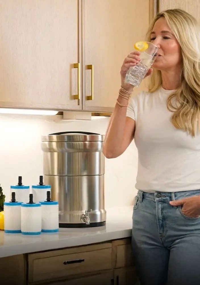 A woman in a white t-shirt drinking a glass of water with a lemon slice in a bright kitchen. On the counter next to her is a stainless steel gravity-fed water filtration system and four replacement filters.