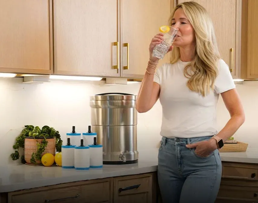 A woman in a white t-shirt drinking a glass of water with a lemon slice in a bright kitchen. On the counter next to her is a stainless steel gravity-fed water filtration system and four replacement filters.