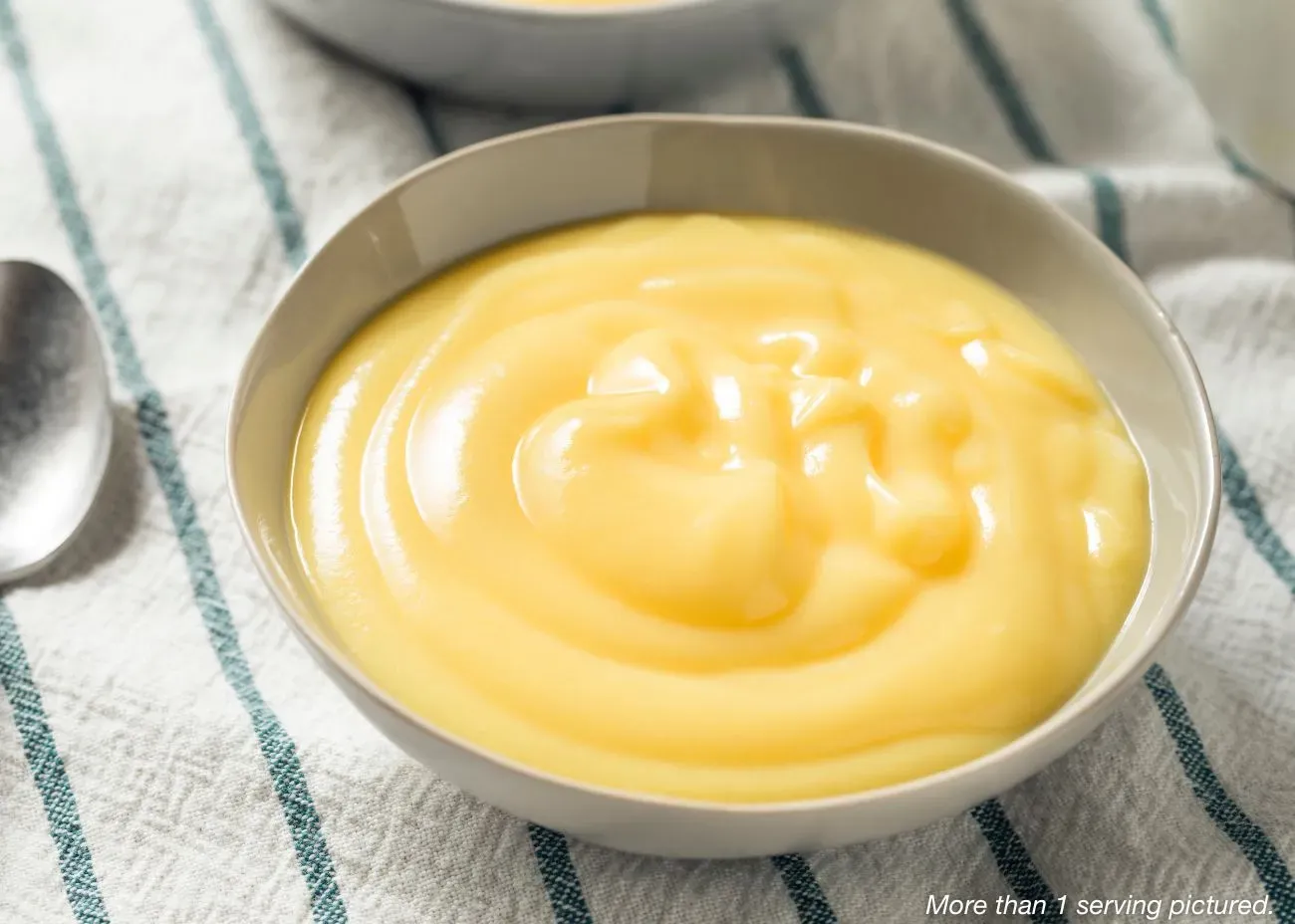 Old-Fashioned Vanilla Pudding A grey ceramic bowl filled with smooth, creamy yellow vanilla pudding, showing soft swirls on the surface, set on a striped linen napkin with a spoon nearby.