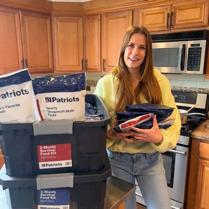 A woman proudly holds emergency food pouches next to two stacked 4Patriots "3-Month Survival Food Kits" on a kitchen counter.