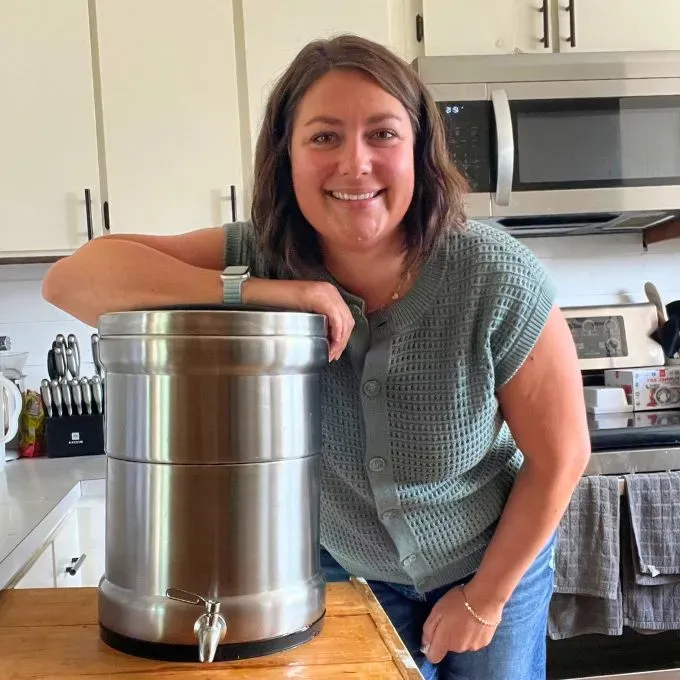 A woman smiles while leaning on a large, stainless steel Ultimate Water Filtration System set on a kitchen counter.