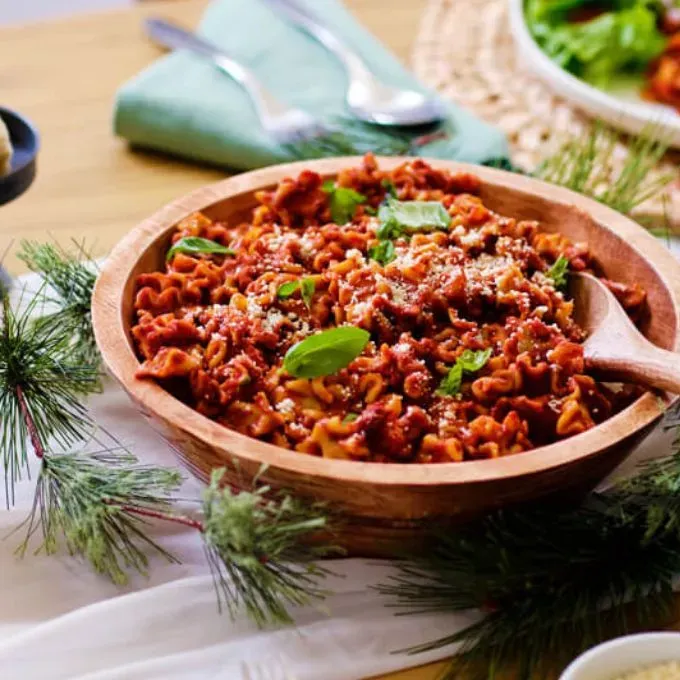 A festive wooden bowl of pasta from the 3-Month Emergency Food Kit, topped with grated cheese and fresh basil, sitting on a holiday-themed table with pine garland.