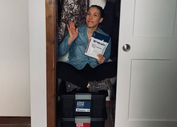 A woman sitting in her closet on top of the 3-Month Survival Food Kit totes while holding a package of the Hearty Stroganoff Style Pasta