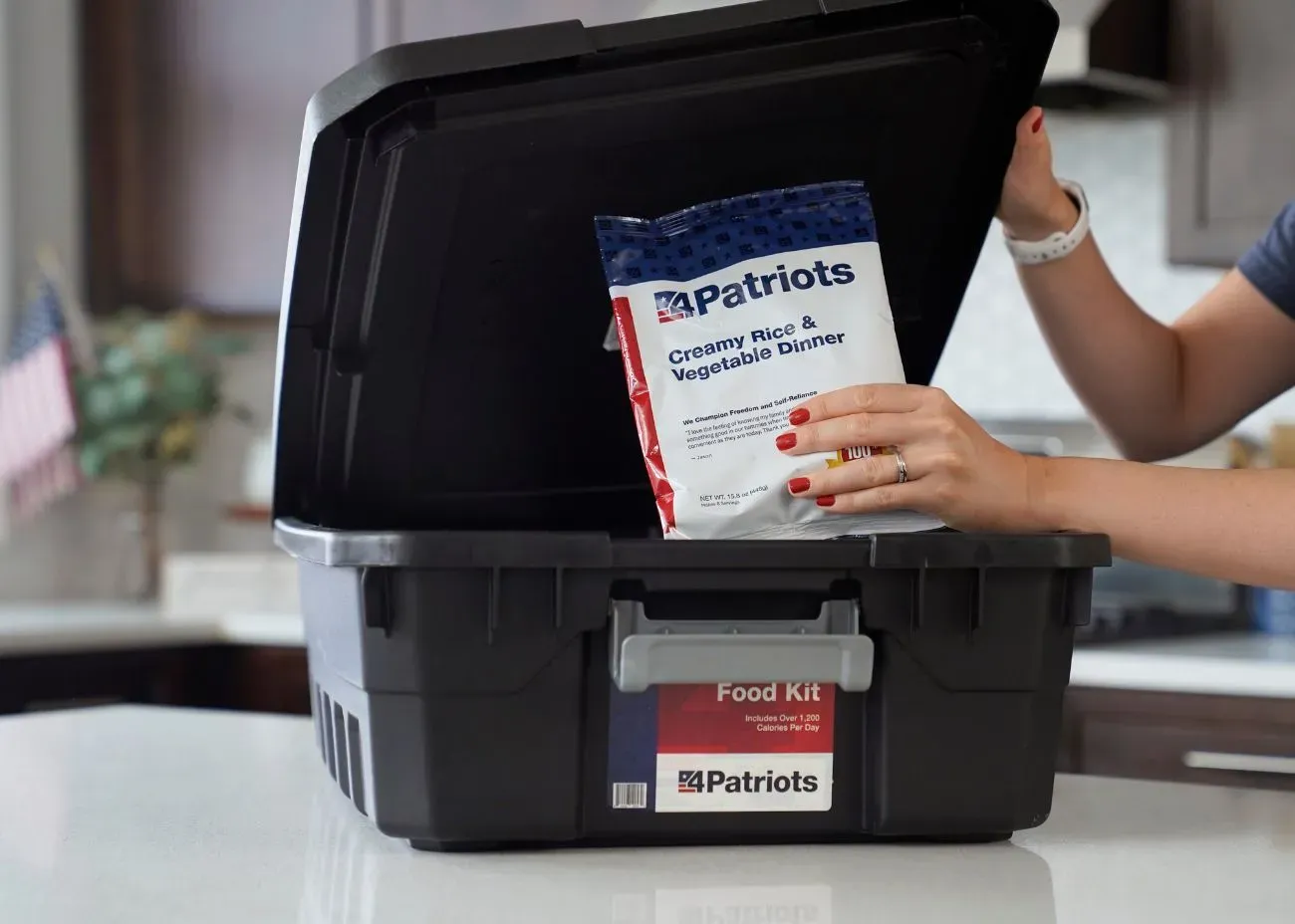 A person placing a 4Patriots Creamy Rice & Vegetable Dinner pouch into a black emergency food supply kit labeled “Food Kit – Includes Over 1,200 Calories Per Day.” The scene is set in a modern kitchen, highlighting preparedness at home.