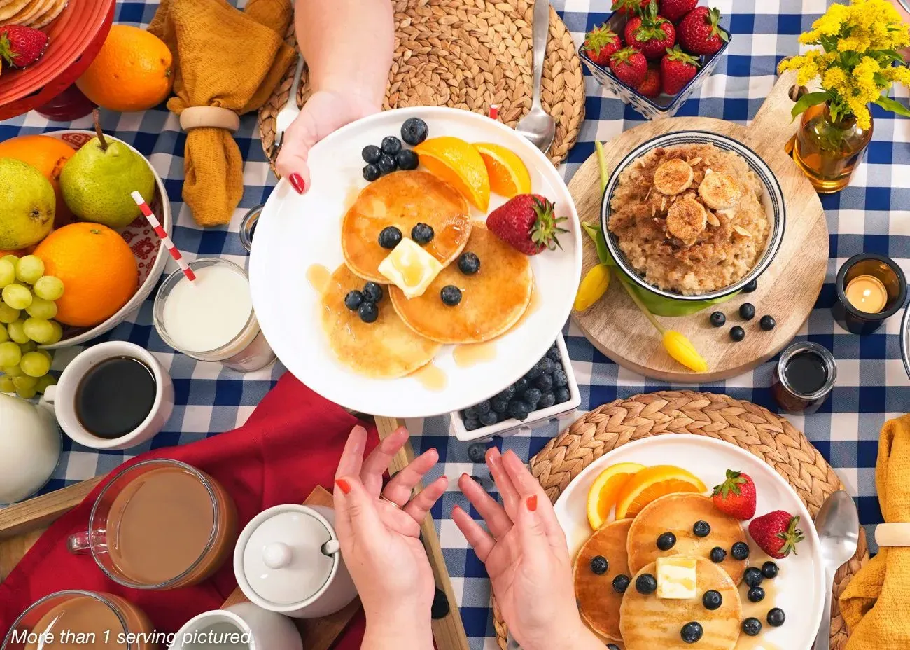 A stack of golden-brown buttermilk pancakes from the 4Patriots 4-Week Emergency Food Supply Kit topped with melting butter and syrup, served on a rustic plate with orange juice and coffee in the background.