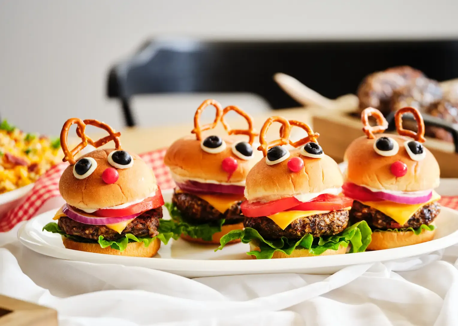 Four 4Patriots Black Bean Burger Mix burgers decorated as reindeer with pretzel antlers, olive eyes, and cherry tomato noses on a white platter.