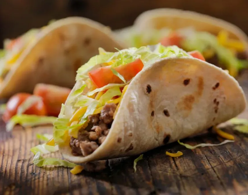 A close-up of a soft taco filled with seasoned ground beef, shredded lettuce, diced tomatoes, and cheese, set on a rustic wooden surface with more tacos in the background.