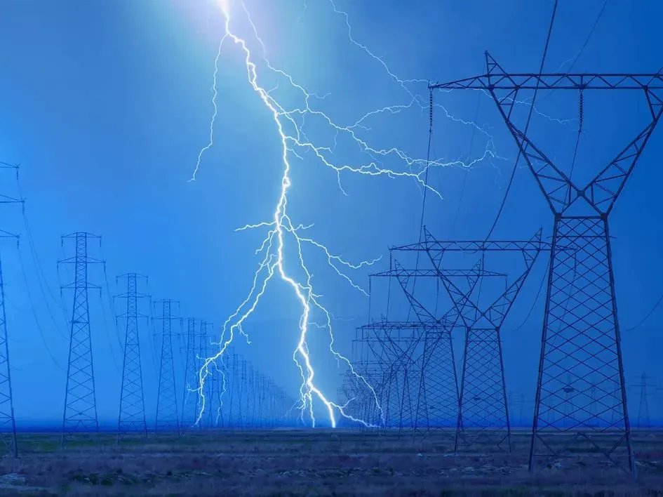 A powerful bolt of lightning strikes the ground between rows of high-voltage transmission towers under a stormy blue sky. This dramatic display highlights the vulnerability of power infrastructure during extreme weather, a critical concern in survival skills and emergency preparedness.