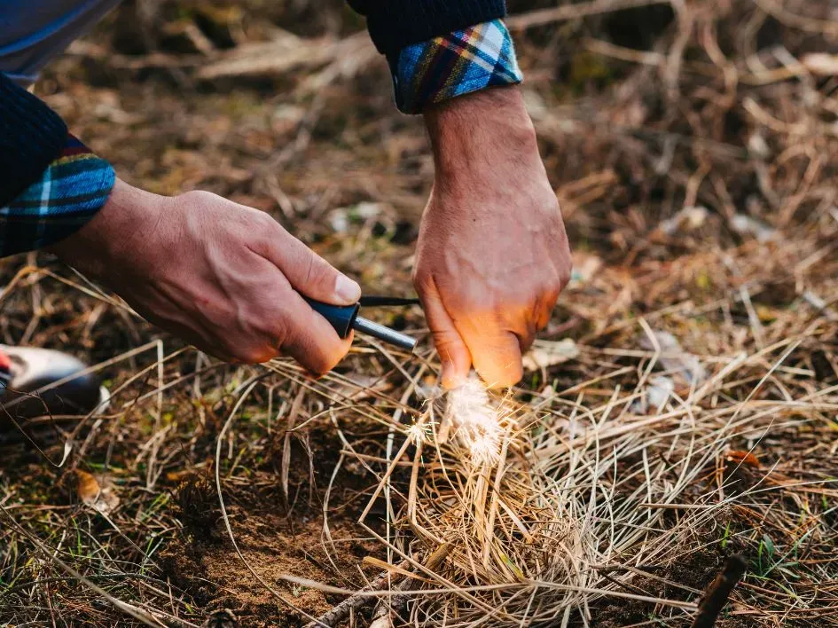 A close-up of a person demonstrating survival skills by using a fire starter to ignite dry grass and twigs on the forest floor. Sparks fly as the tinder begins to catch, showing the essential process of building a fire in the wild.