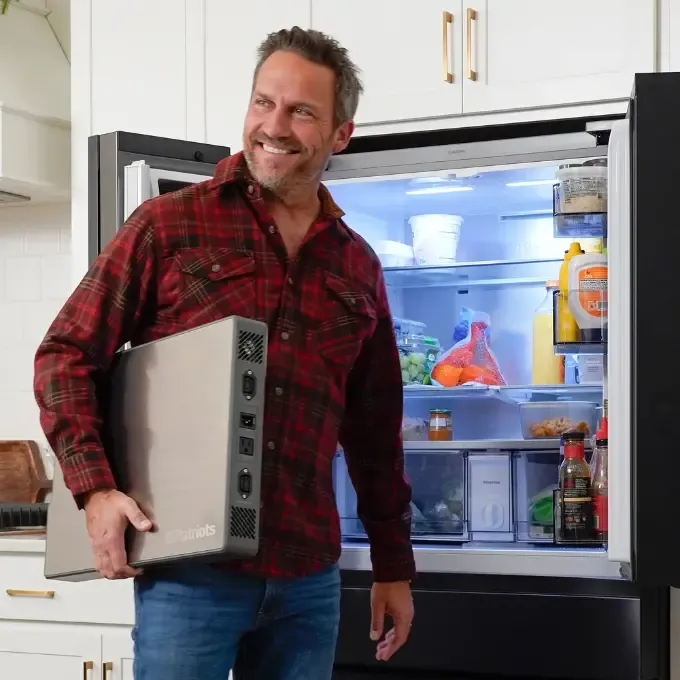 A smiling man in a red plaid shirt holds a 4Patriots Blackout Buddy Emergency Power Supply unit, while standing in front of an open refrigerator filled with food and drinks in a modern kitchen.