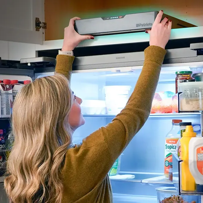 A woman reaches above an open kitchen refrigerator to place a 4Patriots Blackout Buddy Emergency Power Supply on top, while the fridge interior glows with shelves of food and drinks visible inside.