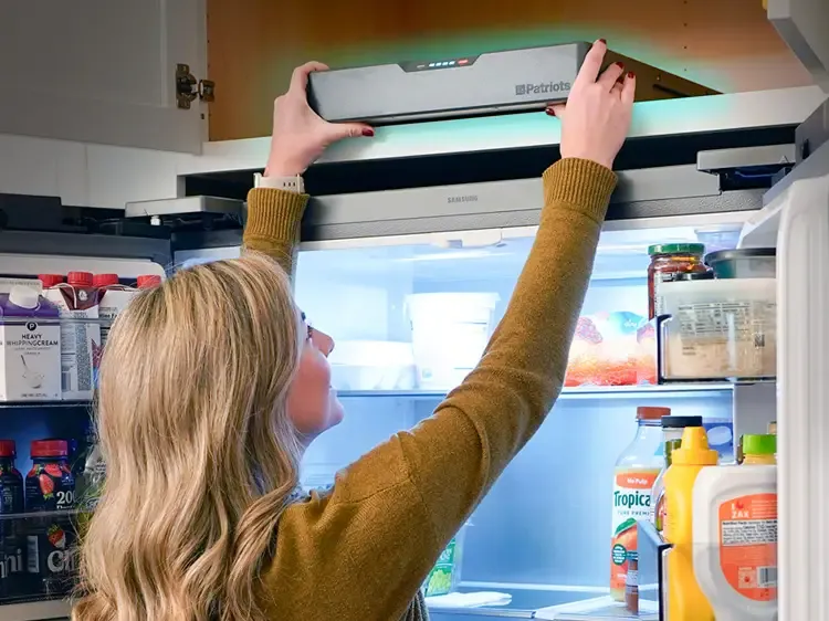 A woman reaches above an open kitchen refrigerator to place a 4Patriots Blackout Buddy Emergency Power Supply on top, while the fridge interior glows with shelves of food and drinks visible inside.