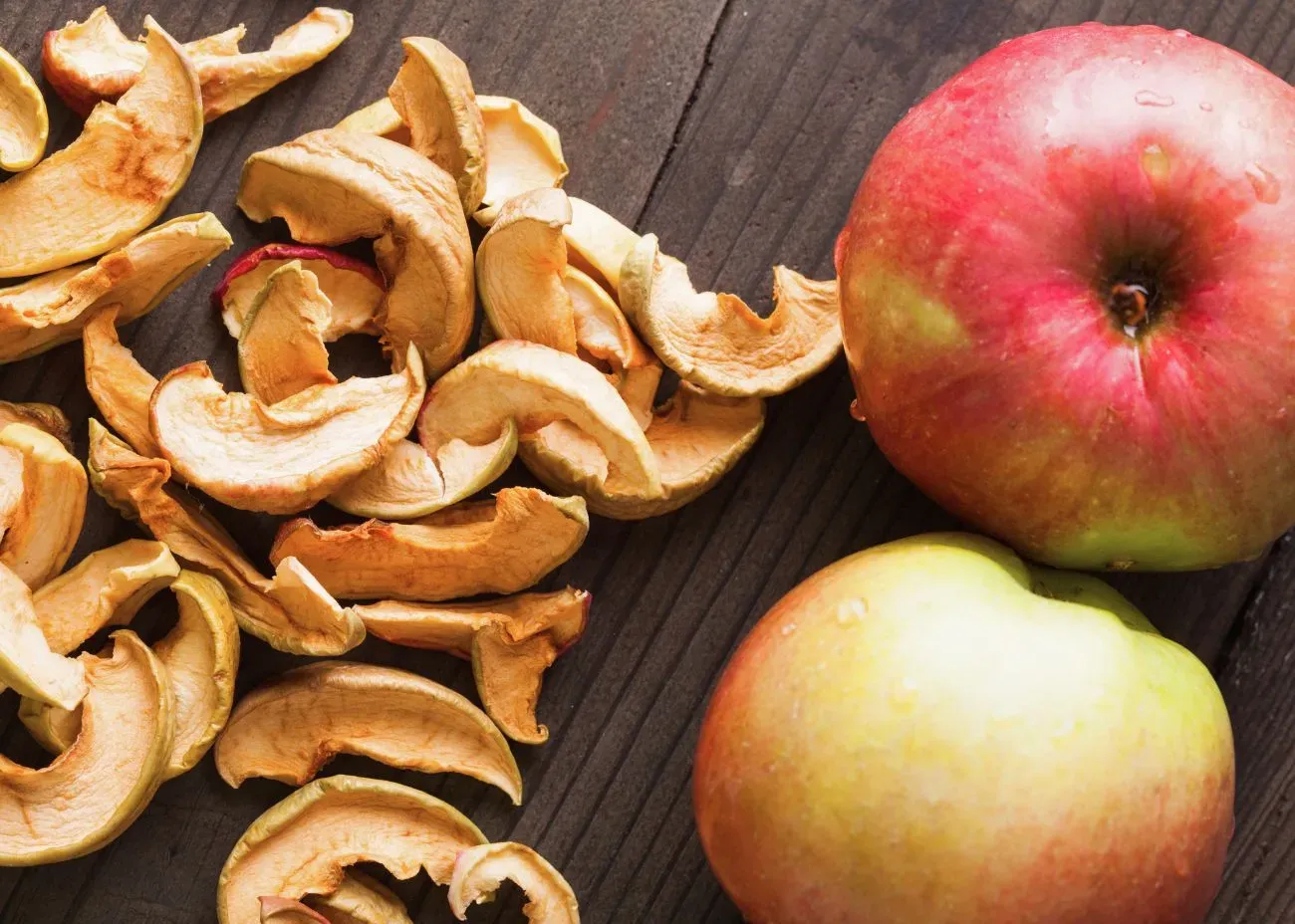 Fresh apples and a pile of dried apple slices arranged on a wooden surface, showing the contrast between juicy fruit and crisp dehydrated pieces.