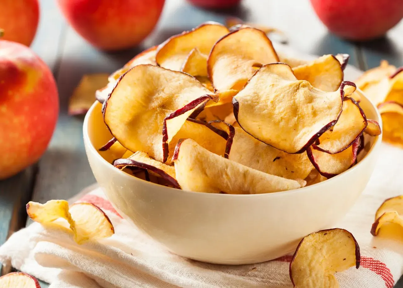 A bowl filled with crispy apple chips, their curled edges and golden color highlighted, with whole apples and scattered chips in the background.