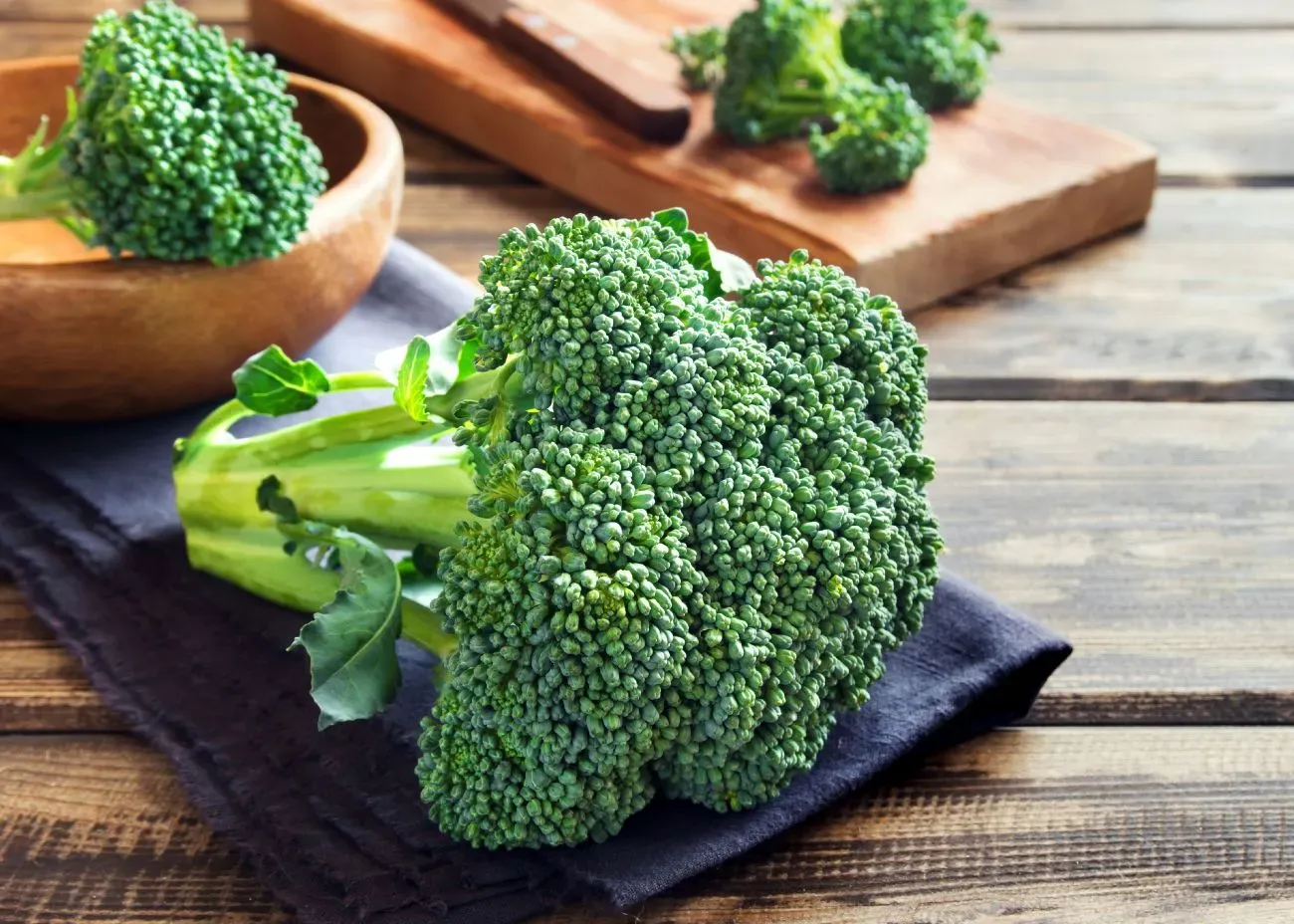 Fresh broccoli heads resting on a dark cloth atop a rustic wooden table, with a wooden bowl and cutting board in the background.