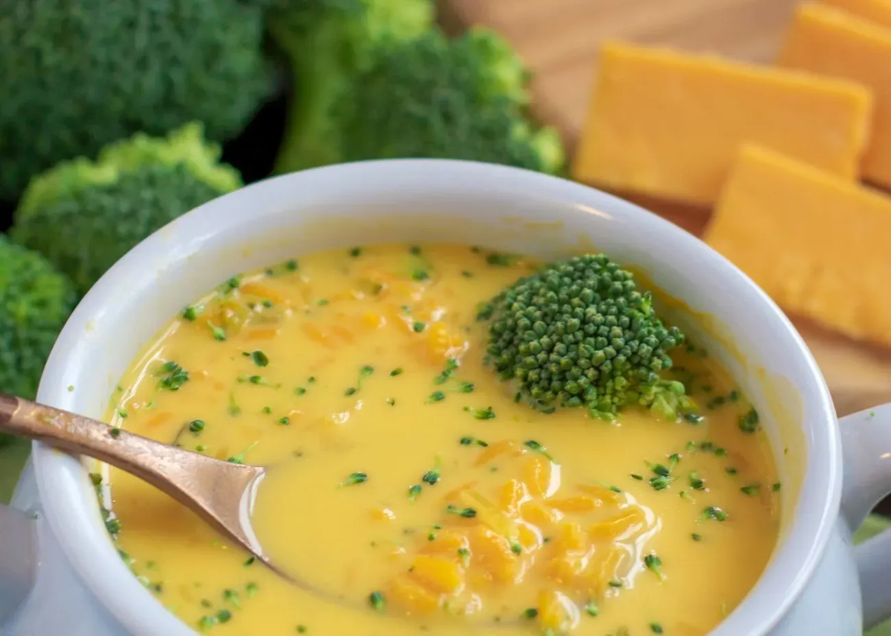 A bowl of creamy broccoli cheddar soup, filled with chunks of broccoli and melted cheese, with fresh broccoli florets and cheddar slices in the background.