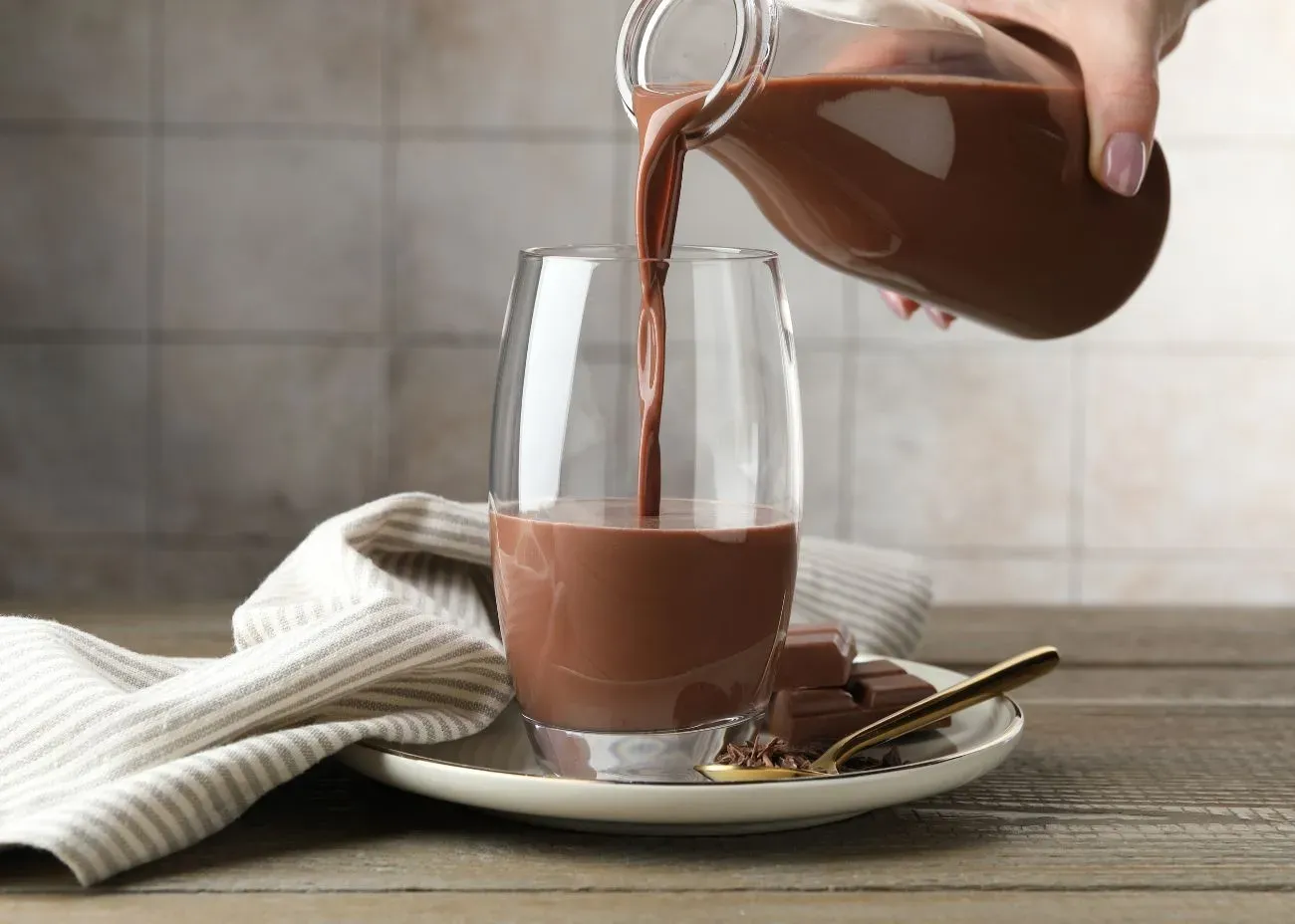 Chocolate milk being poured from a glass bottle into a clear glass, set on a plate with chocolate squares and a gold spoon beside a striped napkin.