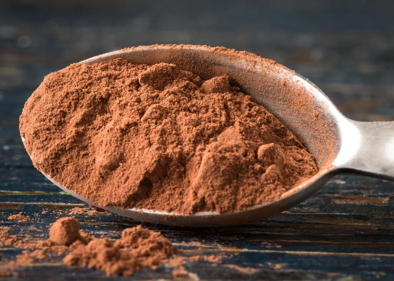 A metal spoon heaped with cocoa powder, with some powder spilled onto a rustic dark wooden surface, highlighting its soft, velvety texture.