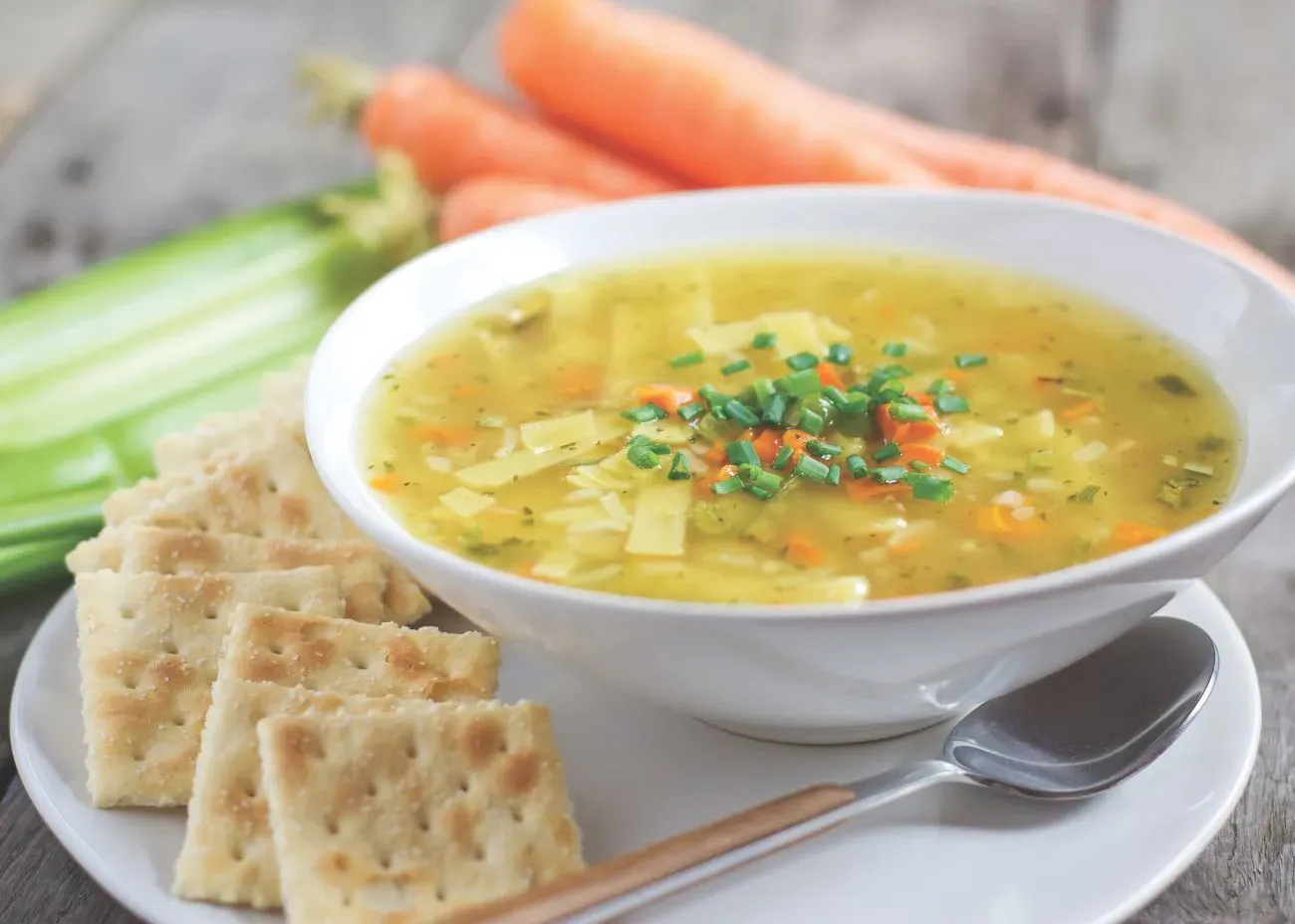 A bowl of golden chicken noodle soup filled with noodles, diced carrots, celery, and herbs, topped with chopped green onions. The bowl is served with saltine crackers on a white plate, with fresh carrots and celery in the background.