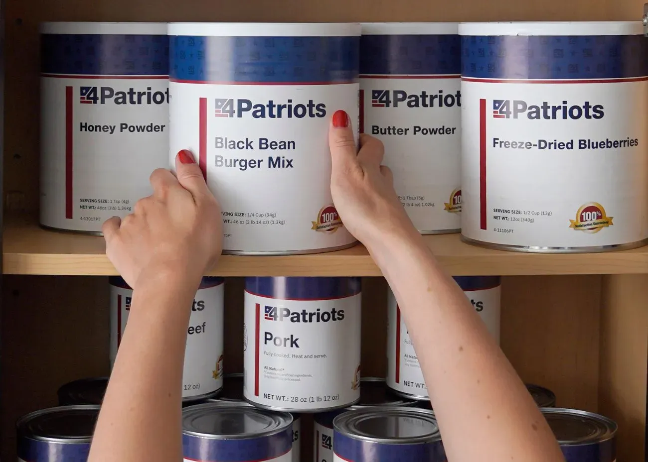 Hands placing a 4Patriots can of Black Bean Burger Mix on a pantry shelf among other emergency food containers labeled Honey Powder, Butter Powder, Freeze-Dried Blueberries, Pork, and Beef.