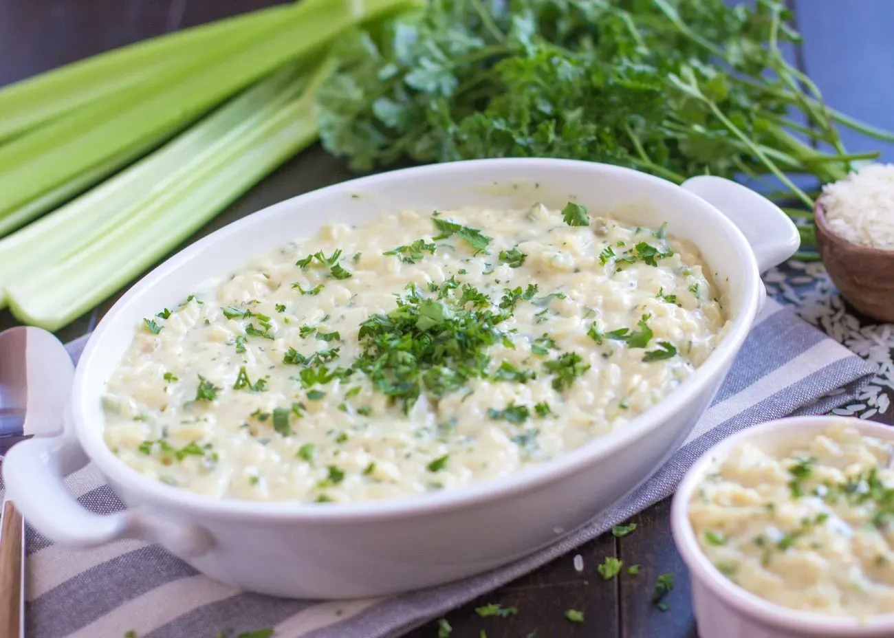A white casserole dish filled with creamy chicken and rice soup garnished with chopped parsley, surrounded by fresh celery stalks and herbs on a striped napkin.