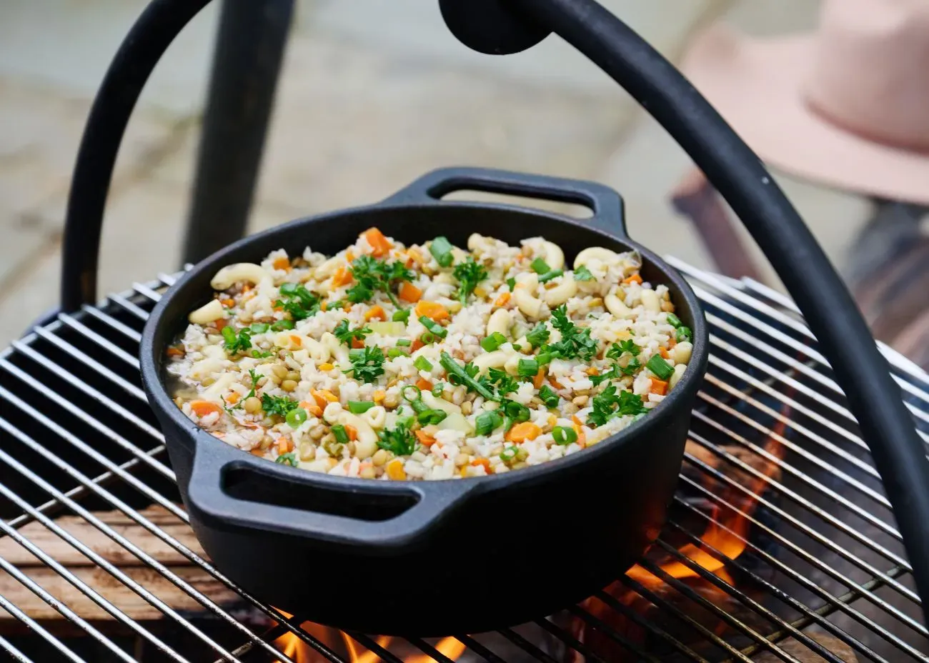 A black cast iron pot filled with a hearty campfire meal of rice, vegetables, and pasta cooks over an open flame on a grill grate.
