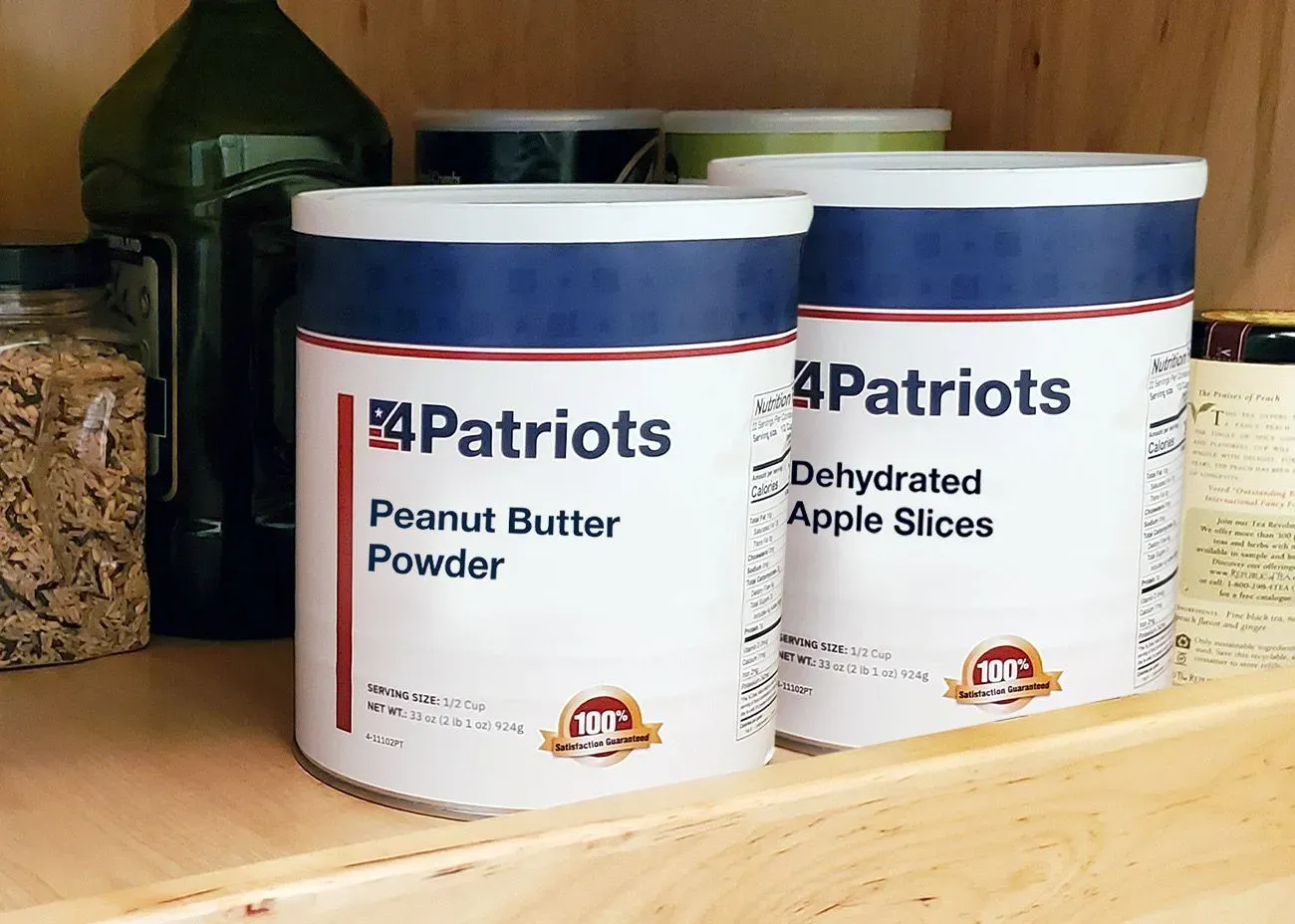 Two large 4Patriots food storage cans on a pantry shelf labeled “Peanut Butter Powder” and “Dehydrated Apple Slices,” surrounded by other pantry items.